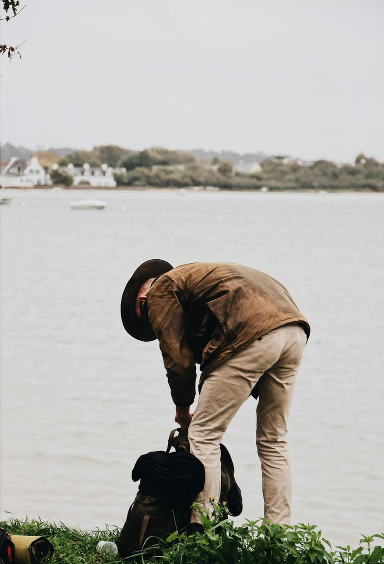 A man in a brown waxed jacket and hat packing a backpack by a scenic lake or river.