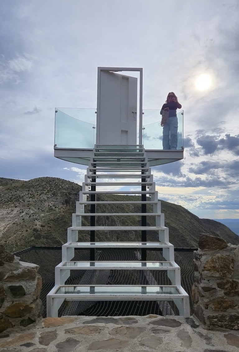 Una mujer se encuentra de pie en una escalera de cristal que conduce a una puerta blanca con vistas a un paisaje montañoso.