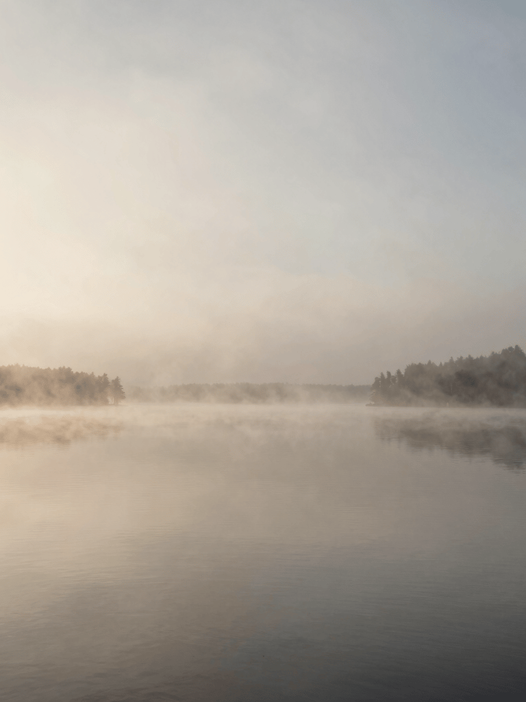 A serene wide-angle landscape photograph of a misty lake in Ontario, Canada, at dawn. The water is perfectly still, reflecting a soft beige and muted grey sky. Minimalist photography style with ample negative space.