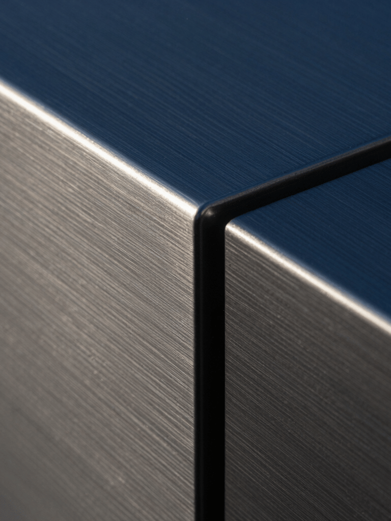 Macro photography of a brushed steel surface with sharp, geometric edges. The lighting creates dramatic shadows in deep blue and charcoal tones, emphasizing a clean and cutting-edge industrial design aesthetic found in high-end North American architecture.