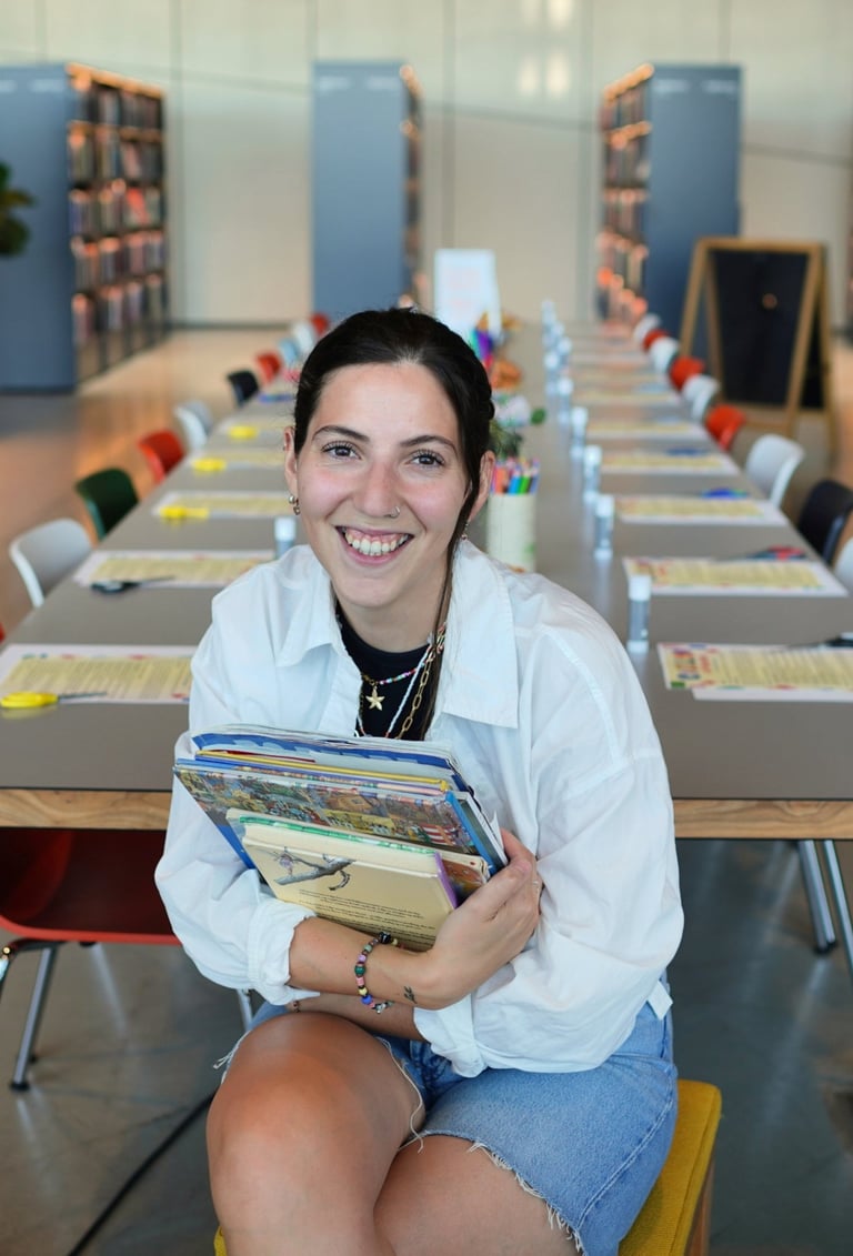 Smiling woman holding a stack of books while sitting in a modern library with study tables.