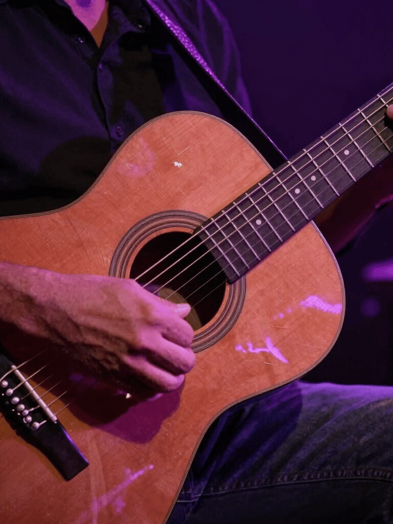 Close-up of a musician's hands playing an acoustic guitar with intensity, shot in a dimly lit South American venue with deep purple and rose colored ambient lighting reflecting on the wood.