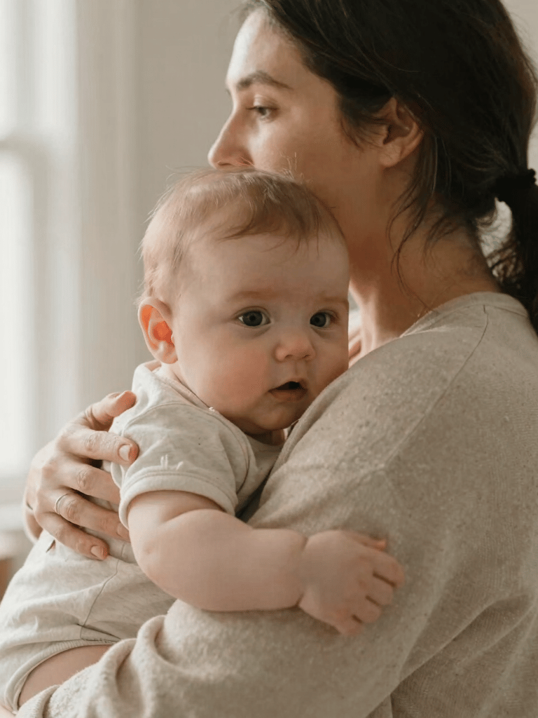 A close-up, high-key photograph of a mother embracing her baby. Soft morning light fills the room, highlighting beige and taupe textures. North American / US (West Michigan) setting.