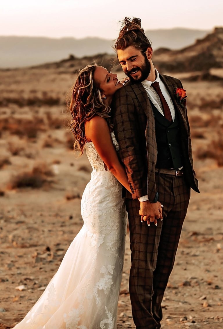 Bride and groom standing together in a desert landscape at sunset, smiling and holding hands