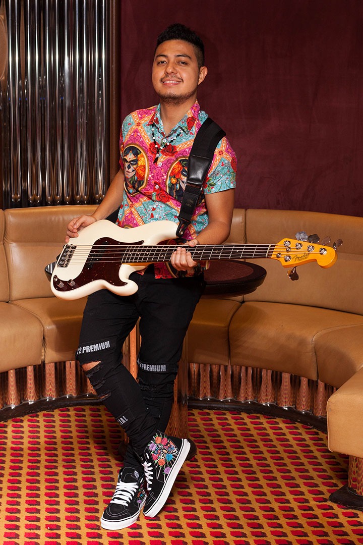 Bassist Ian Yoshio leaning against a coffee table, wearing a colorful shirt, with his white Fender bass guitar.