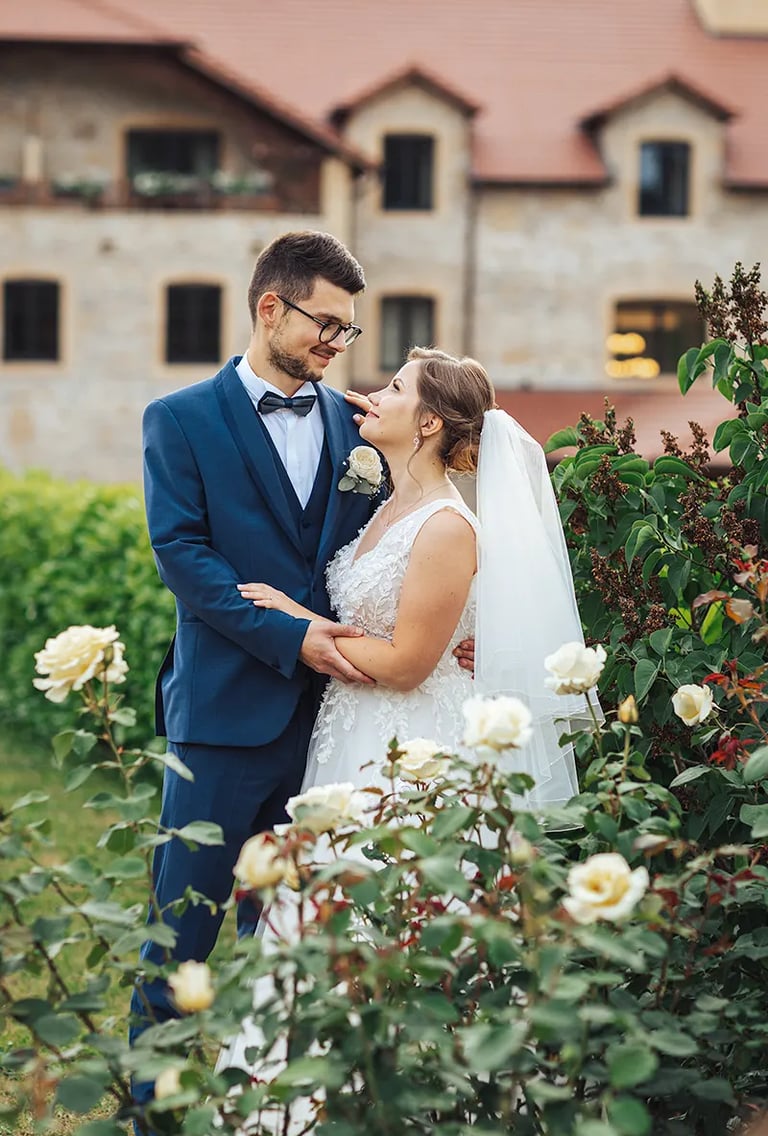 Couple de Mariés qui se regardent tendrement derrière un buisson de roses blanches.