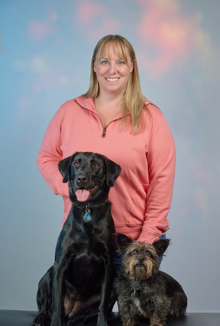 A smiling woman in a pink sweatshirt posing for a portrait with her black Labrador and small terrier mix dog.