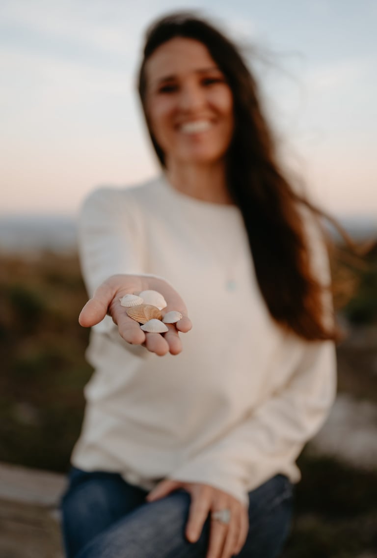 Michael Alexa Pitts smiling, sitting on a boardwalk railing over sandunes while extending a handful of white seashells to you