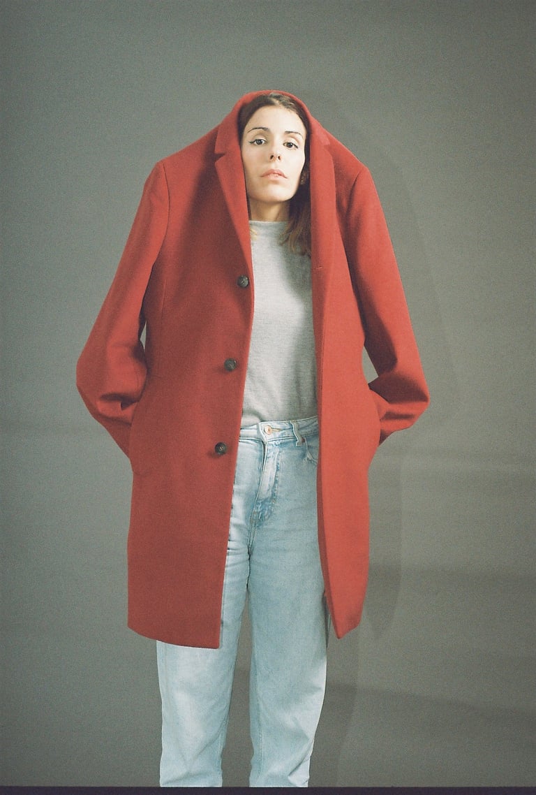 A woman wearing a large red wool coat over her head with light blue jeans against a grey background.