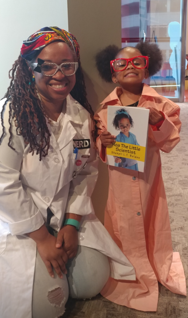 Shenell poses for a pic kneeling in her lab coat with a mini scientist holding her book
