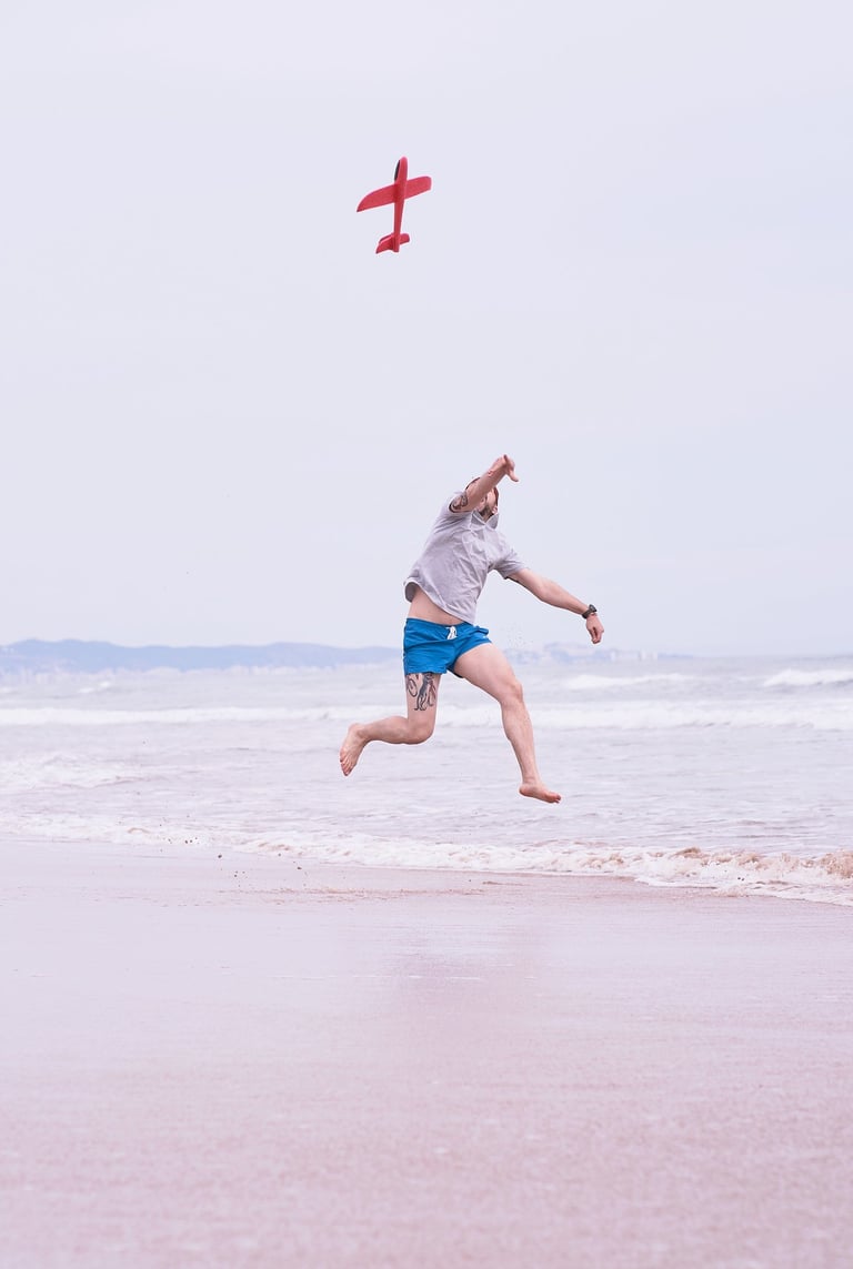 A man jumps on a sandy beach while throwing a red toy glider airplane into the air.