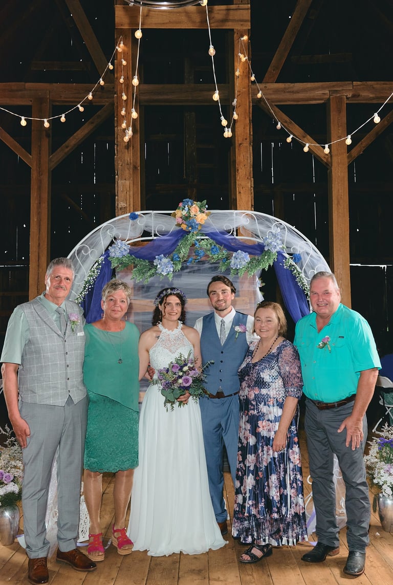 a family wedding picture with a bride and groom in front of wedding arch in a barn