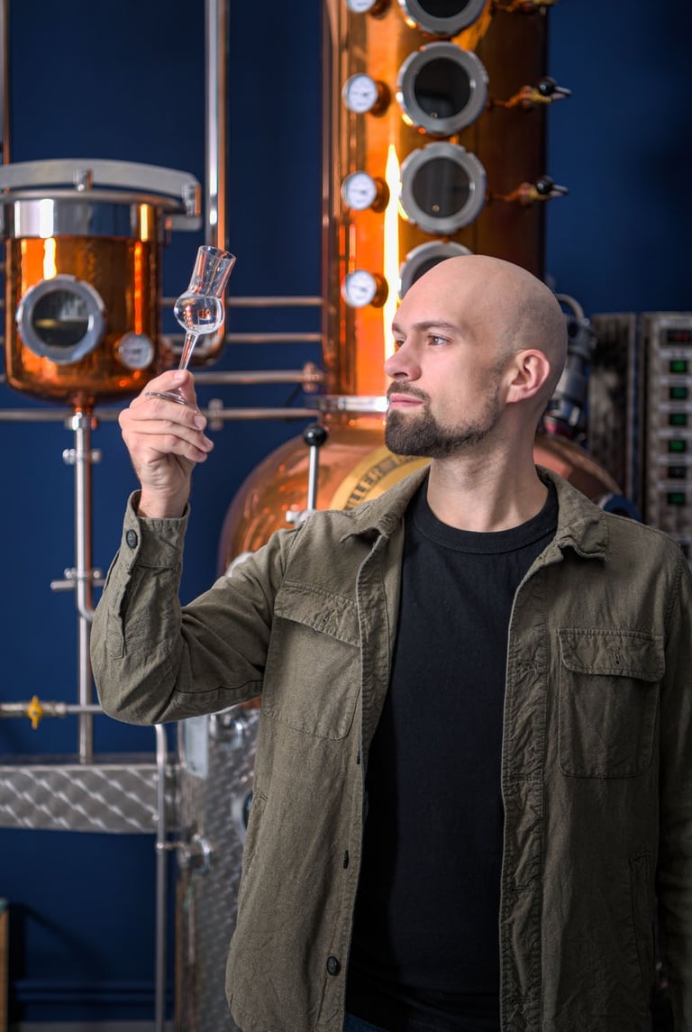mastro distillatore che osserva del liquido in un bicchiere da degustazione davanti a un alambicco