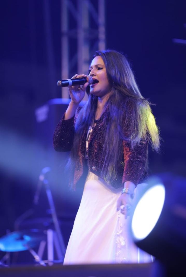 Female vocalist with long hair singing into a microphone during a live concert performance with stage lighting.