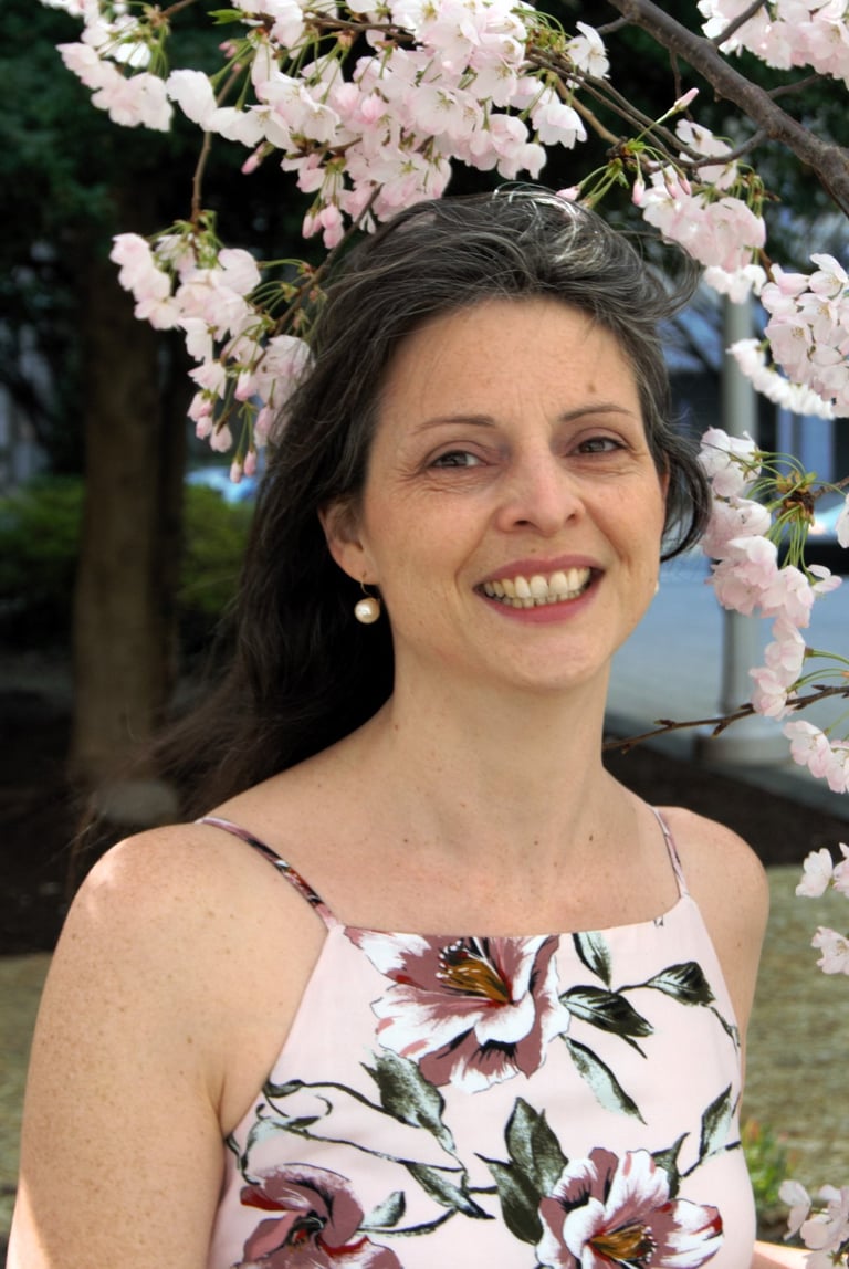 Fair skinned, middle-aged woman with salt and pepper hair, wearing a floral print summer dress against cherry blossom scene