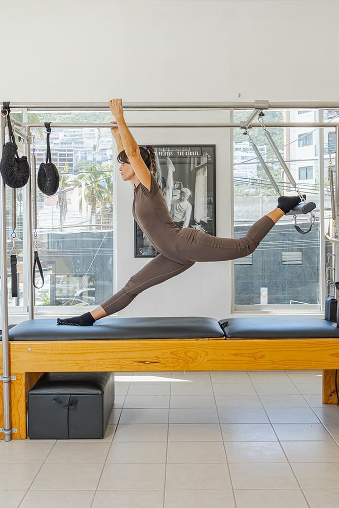 Woman doing a split stretch on a Pilates Cadillac apparatus in a bright studio in Cabo San Lucas.