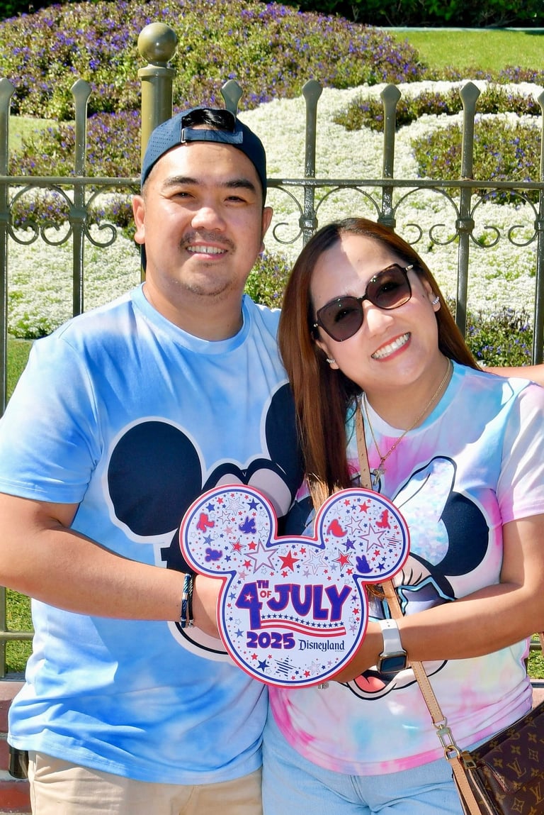 Couple smiling at Disneyland wearing Mickey Mouse shirts and holding a 4th of July 2025 sign.