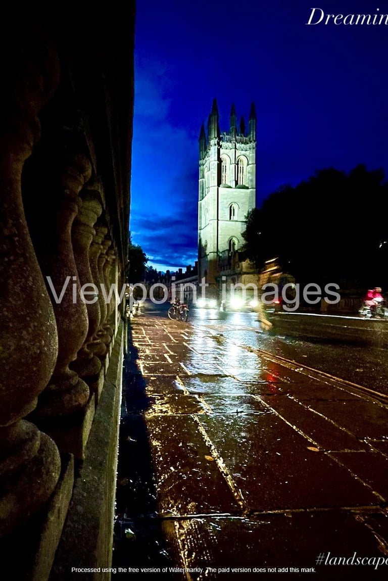 Magdalen College Tower at night viewed from a rainy Oxford street with reflections on wet pavement.