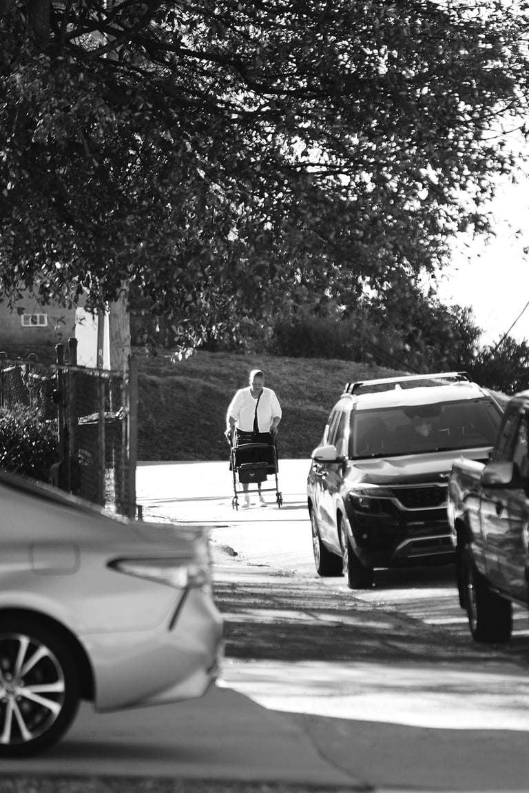 Black and white street photography of an elderly person using a mobility walker on a sidewalk lined with parked cars.