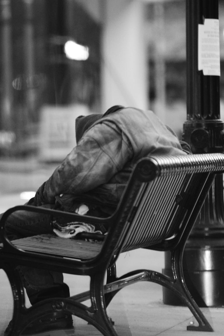 Black and white photo of a homeless person sleeping on a city park bench at night.
