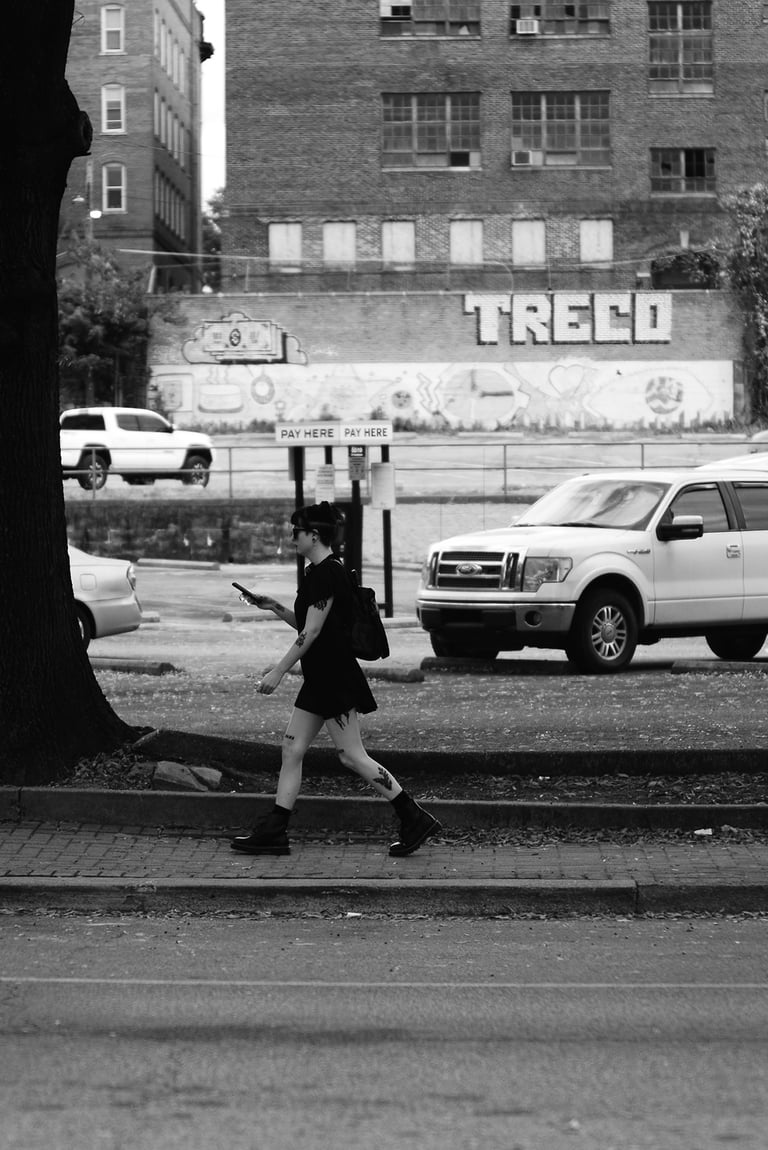 A person walks on a city sidewalk past parked cars, a large tree, and a building with "TRECO" painted on it, in black and
