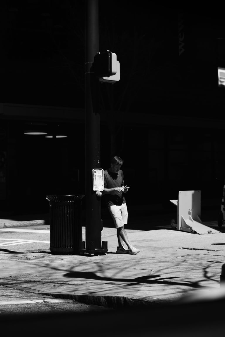 A man leaning against a black lamp post on a city street while using his smartphone in black and white.