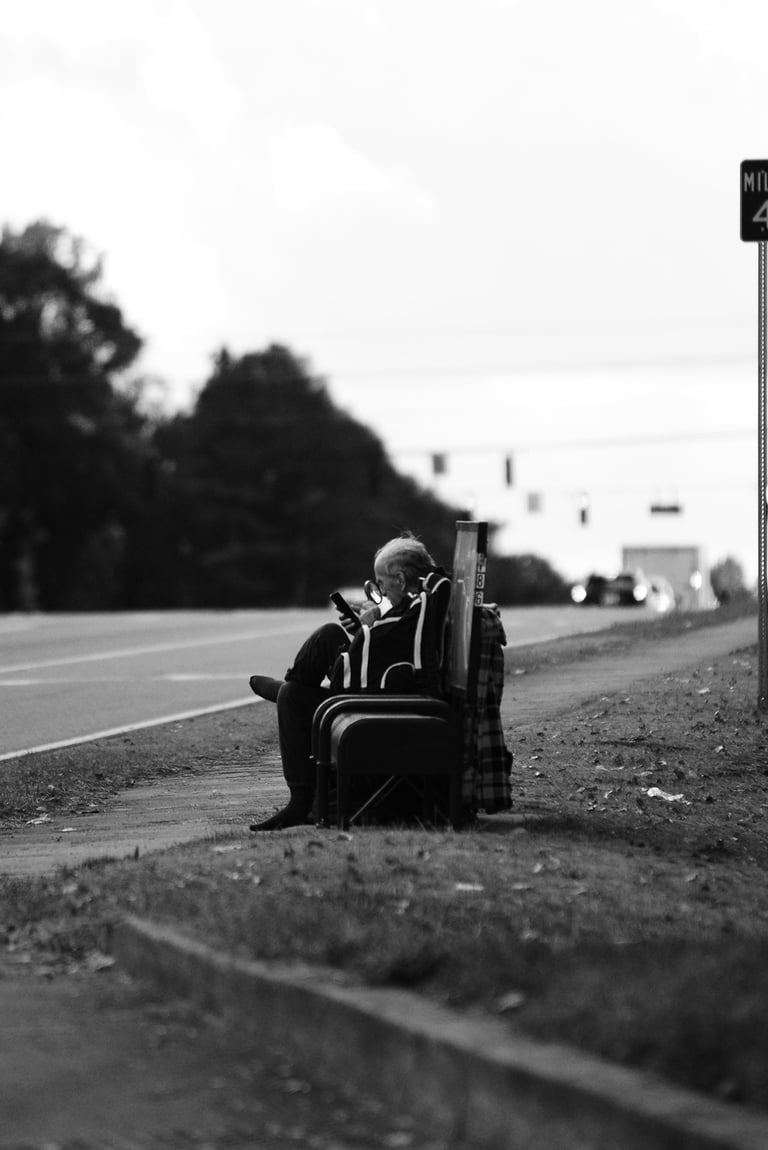 Black and white street photography of a person sitting on a bench by a highway using a smartphone.