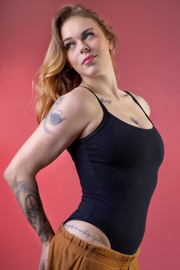 mulher loira de maiô preto e tatuagens a mostra em pose para ensaio sensual com fundo vermelho