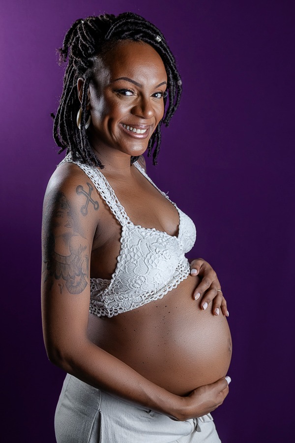 mulher preta gestante com blusa crochê segurando a barriga e sorrindo em fundo roxo