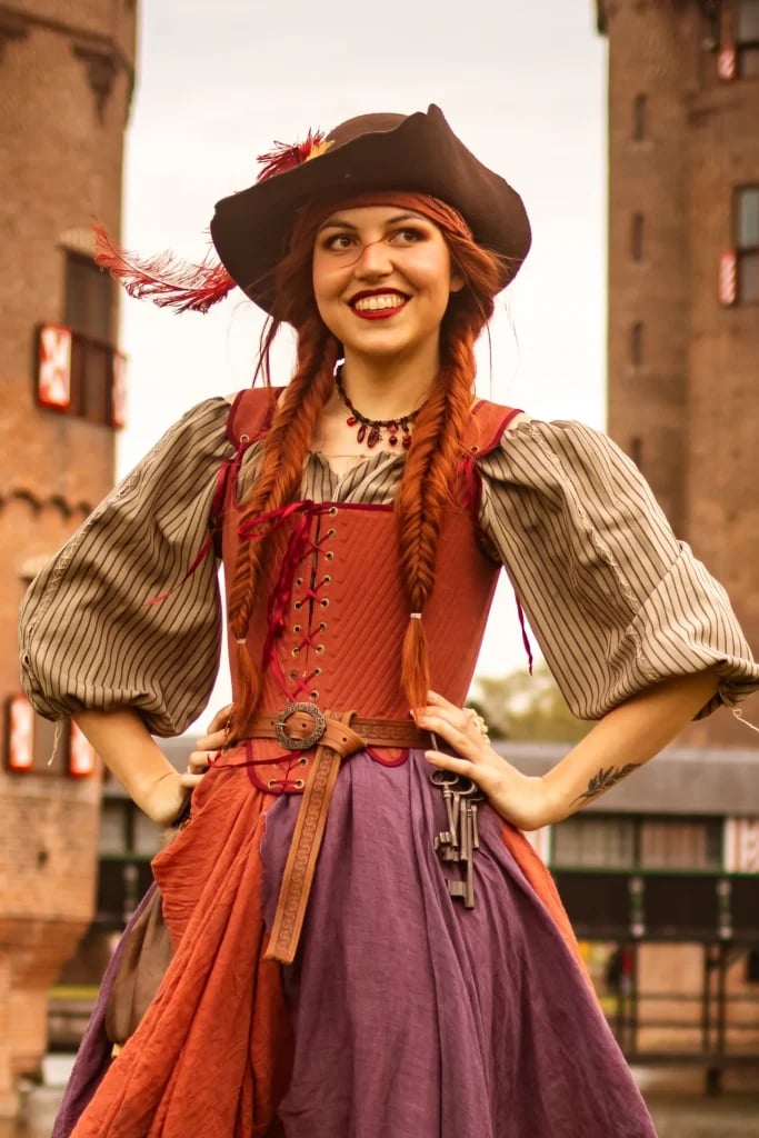 Een vrolijke piratenprinses met een hoed, rode vlechten en een korset poseert op een evenement.