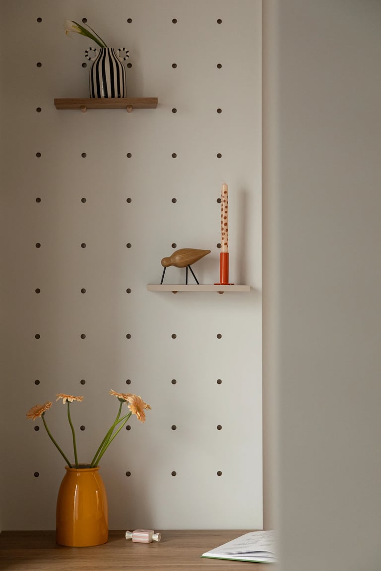 Drewniane biurko, żółty wazon i biała ścianka pegboard z półkami.