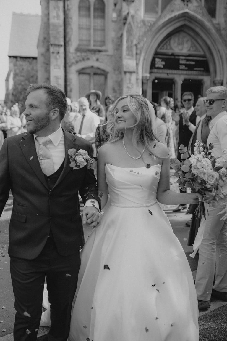 Black and white photo of a bride and groom walking out of a church while guests toss wedding confetti.