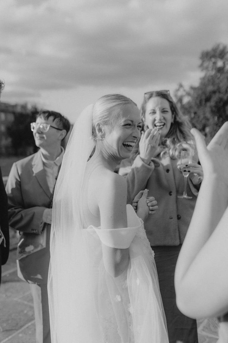 A joyful blonde bride laughs during her outdoor wedding reception with guests in a candid black and white photo.