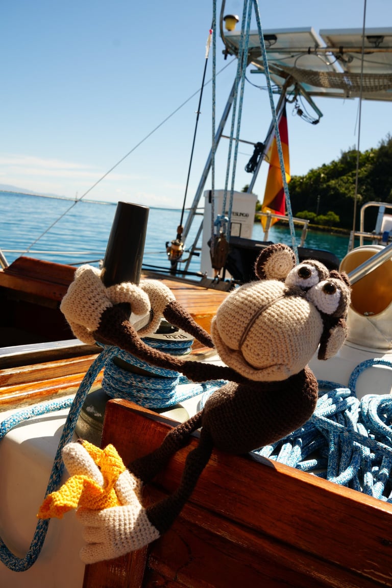 A cute crochet monkey toy holding a winch and banana on a sailboat deck under a clear blue sky.