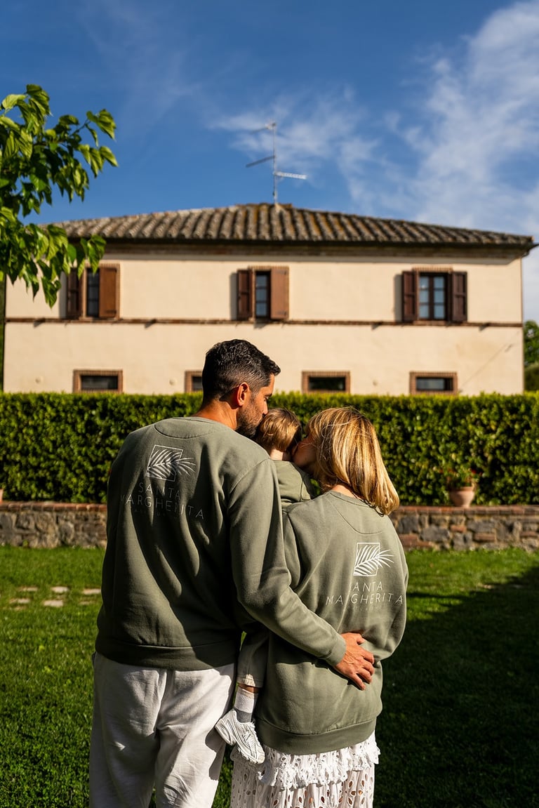 A family wearing matching green sweatshirts hugs in a garden in front of a rustic Italian villa.