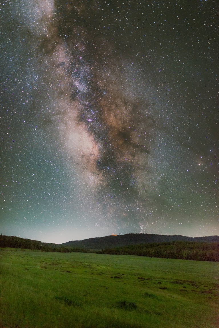 Milky Way galaxy stretching across a starry night sky above a dark forest and green meadow.