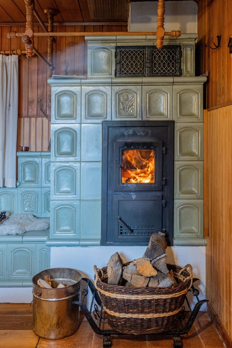 Traditional ceramic tile stove with a roaring fire and firewood basket in a cozy wood-paneled room.