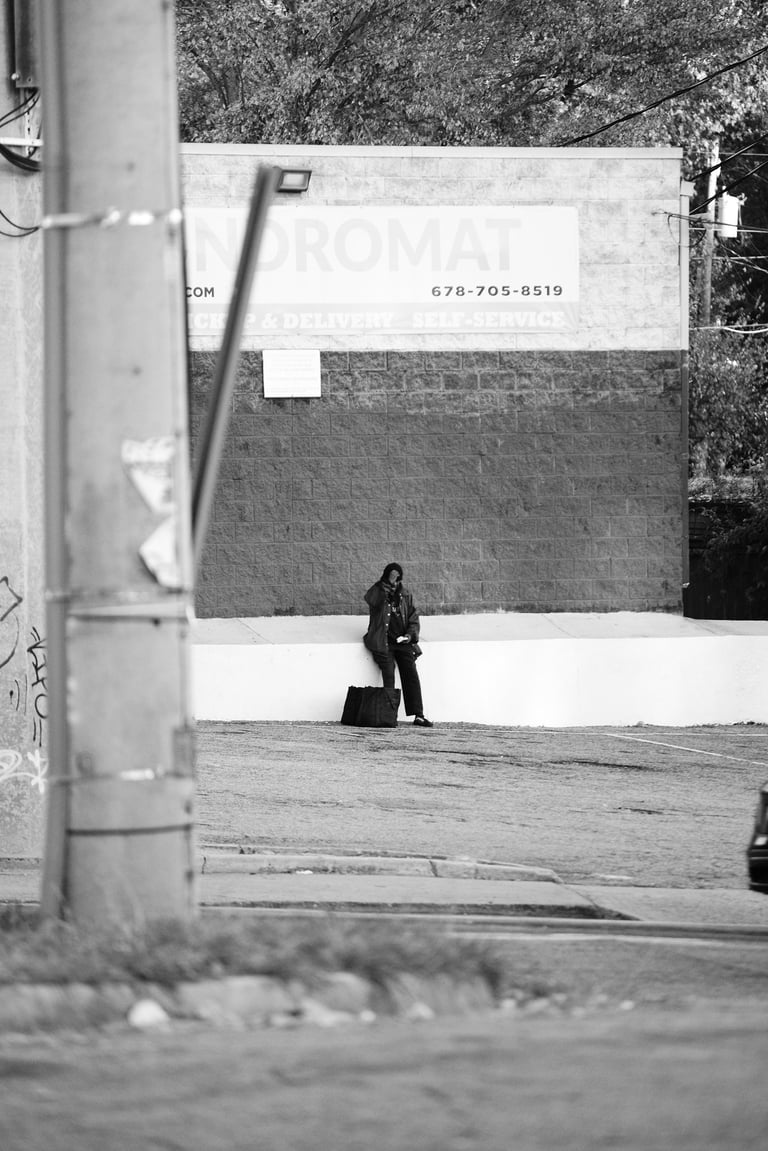 Black and white street photography of a person sitting outside a laundromat in an urban area.