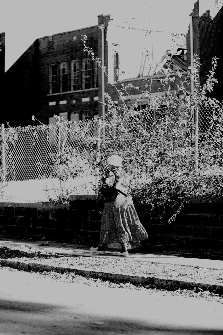 A black and white street photography shot of a woman walking past a chain-link fence and ruined building.