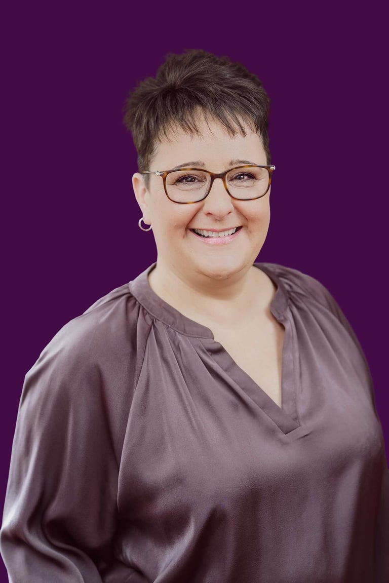 Professional corporate headshot of a smiling woman with glasses and short hair against a purple background.