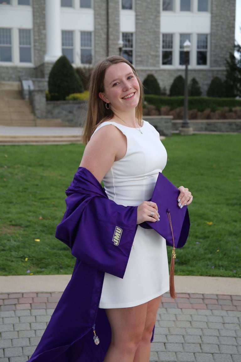 Smiling JMU graduate in a white dress holding a purple graduation cap and gown on campus.