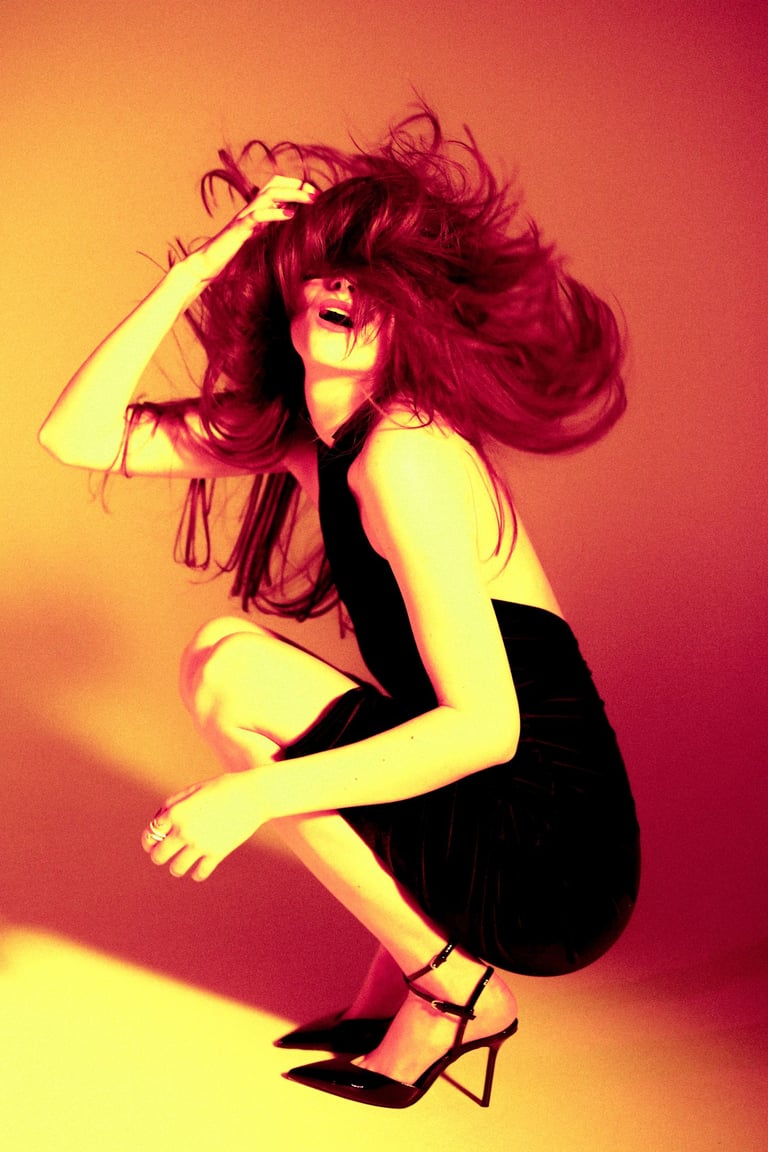 Fashion model in a black mini dress and high heels posing with wind-swept hair under warm orange lighting.