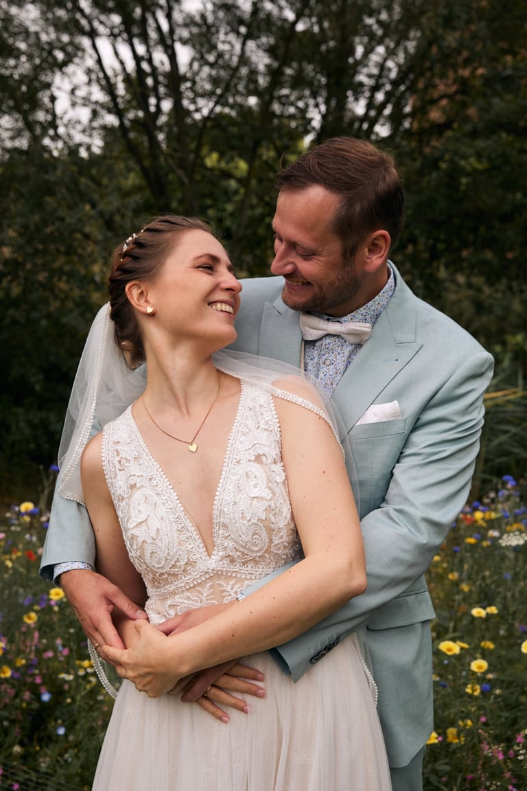 Smiling bride in lace wedding dress and groom in light blue suit posing in a wildflower garden.