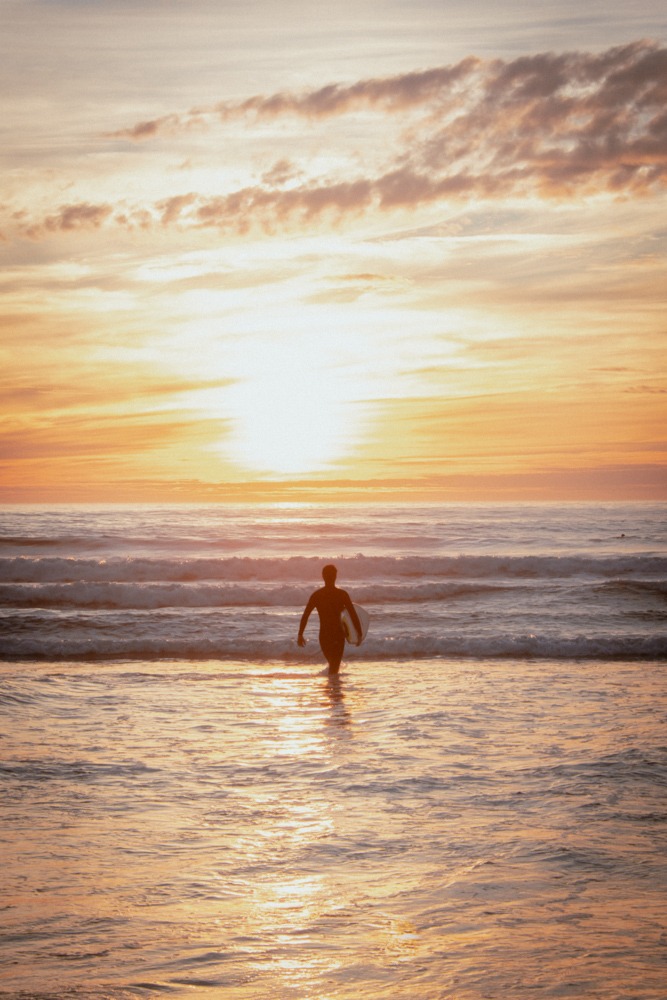 A silhouette of a surfer standing in the ocean, walking into the sun. Vibrant orange and yellow