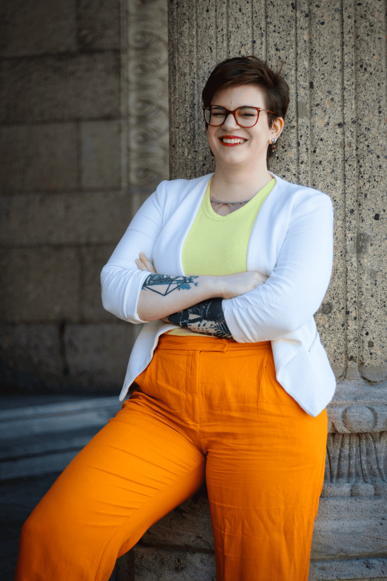 ein Foto von Swana Schuchmann. Sie trägt einen weißen Blazer und lehnt sich an eine Säule, lachend.