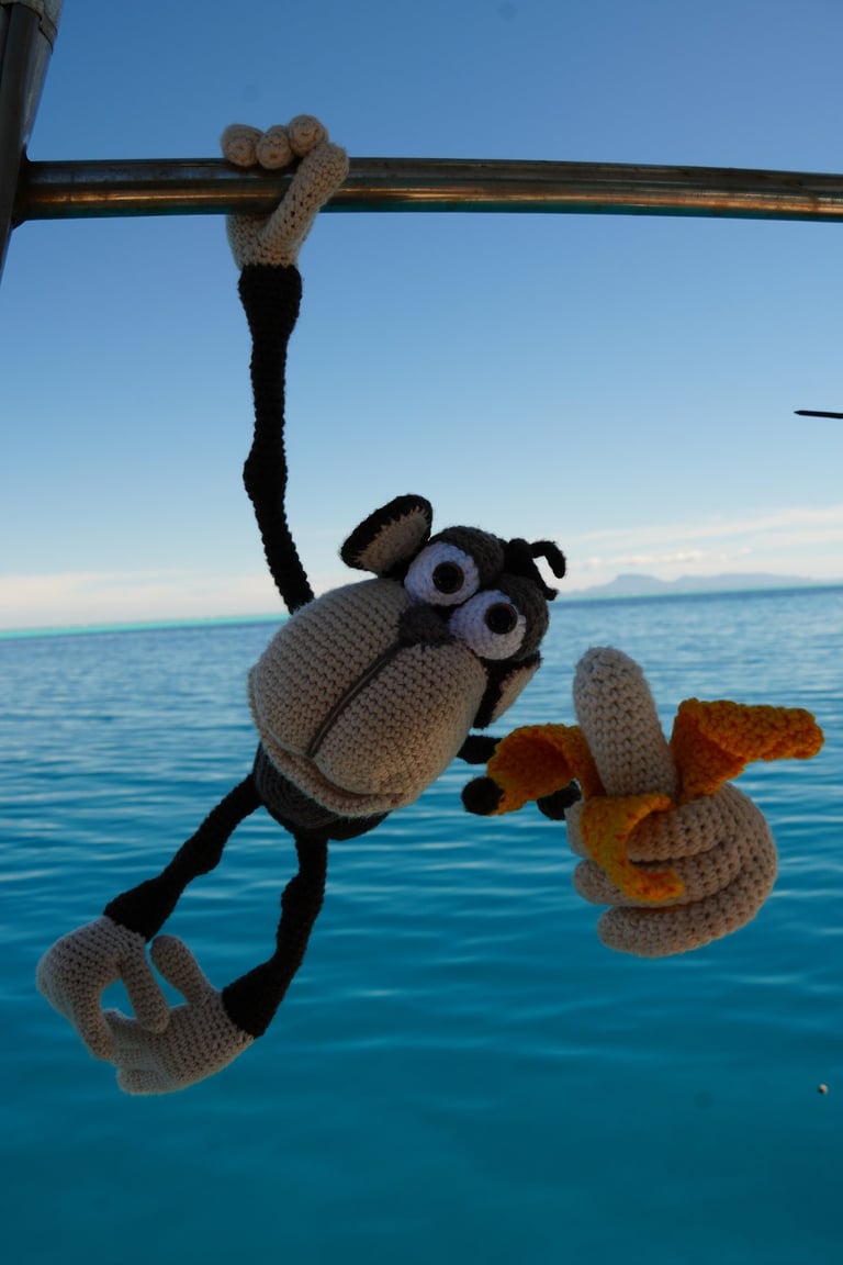 A crochet chimpanzee hanging from a boat rail holding a yarn banana over blue tropical ocean water.