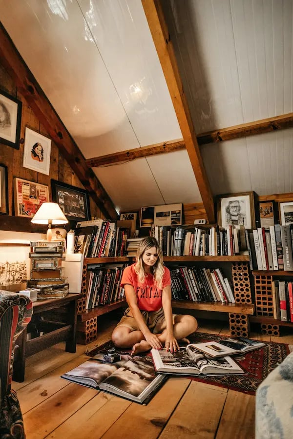 A woman sits on the floor of a cozy attic home library reading books near wooden shelves.