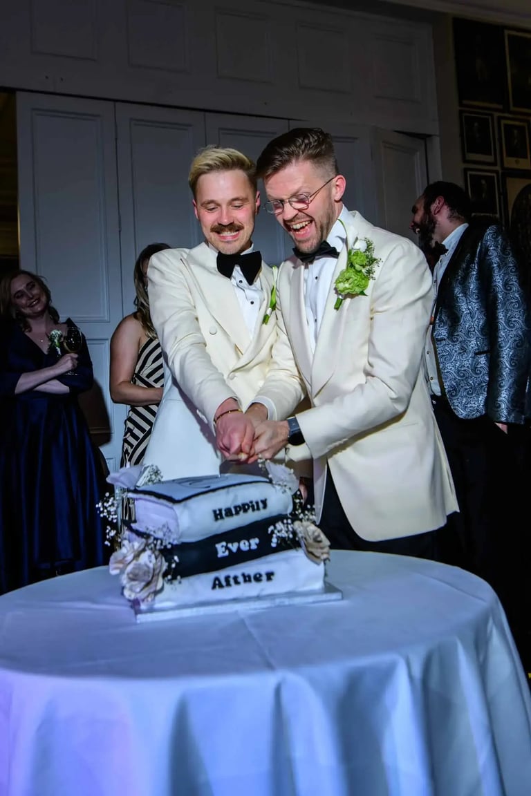 A happy groom couple in white tuxedos cutting their black and white tiered wedding cake.