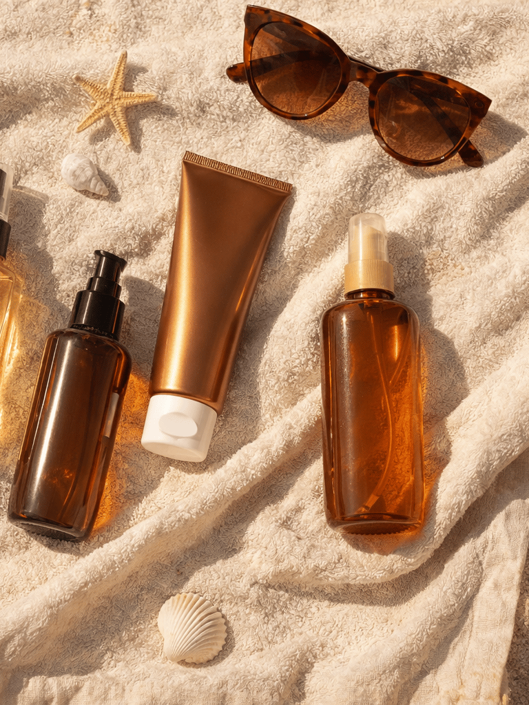 Sunscreen bottles, tanning oil, and sunglasses on a beach towel with shells.