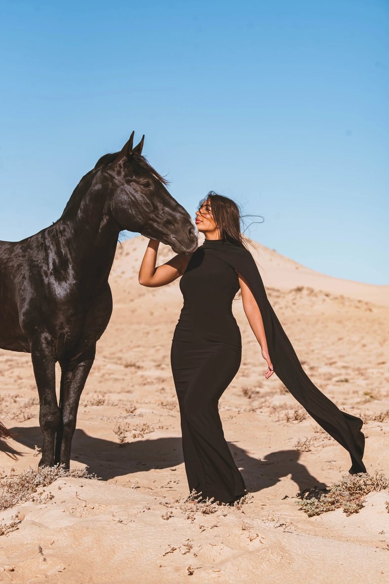 Luxury equestrian fashion shoot in Dakhla featuring a model in a black gown posing with a black horse in the dunes.